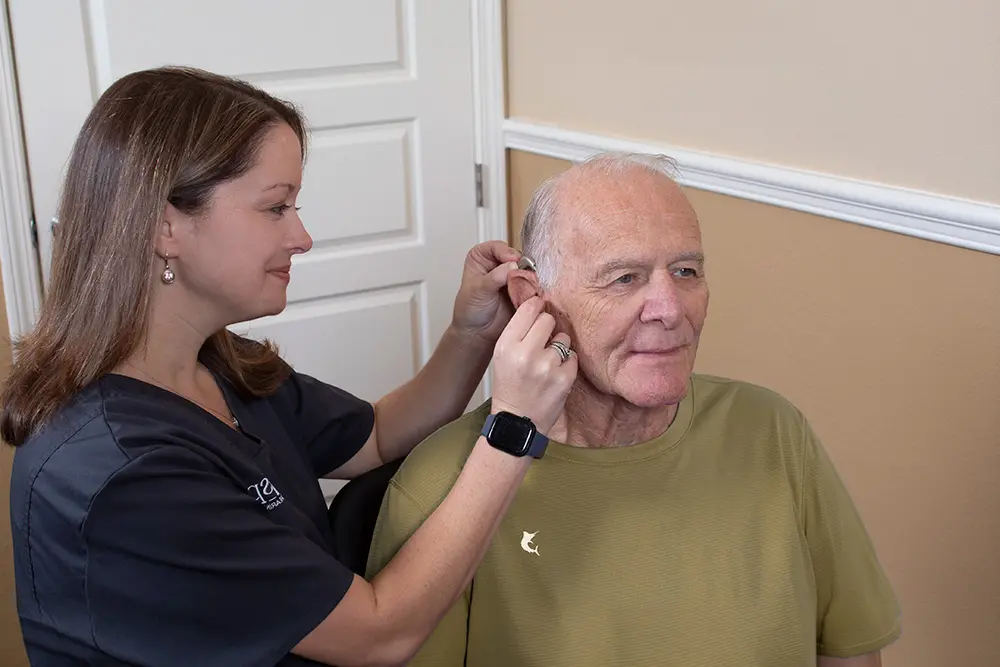 A female healthcare professional helps an older man put on a hearing aid while he sits and smiles in a room with beige walls and a white door.