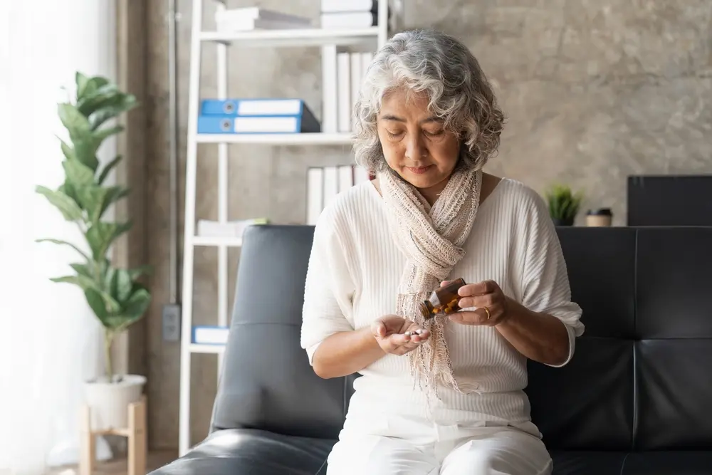 An older woman with gray hair sits on a sofa, looking down as she pours pills from a medicine bottle into her hand. She wears a light scarf and white clothes, with shelves and a plant in the background.