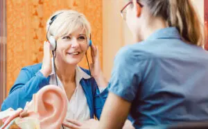 A smiling older woman wearing headphones sits across from a professional during a hearing test, with a large ear model visible on the table between them.