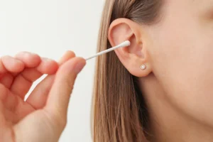 A close-up of a person using a cotton swab to clean the inside of their ear. The person has straight light brown hair and is wearing a small, round earring. The background is plain and light-colored.