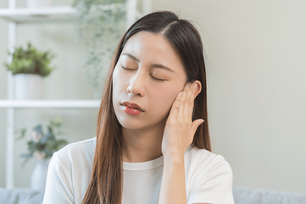 A woman with long hair and closed eyes touches the side of her face near her ear, appearing to be in discomfort. She is indoors, and there are blurred plants in the background.