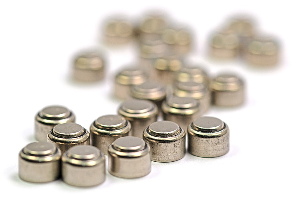 Close-up of several small, round, silver button cell batteries, with some batteries in focus in the foreground and others blurred in the background on a white surface.