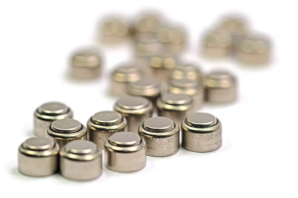 Close-up of several small, round, silver button cell batteries, with some batteries in focus in the foreground and others blurred in the background on a white surface.