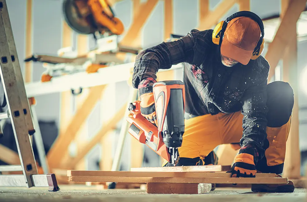 A construction worker wearing safety gear uses a nail gun on a wooden plank at a building site. Wood beams, tools, and construction materials are visible in the background.