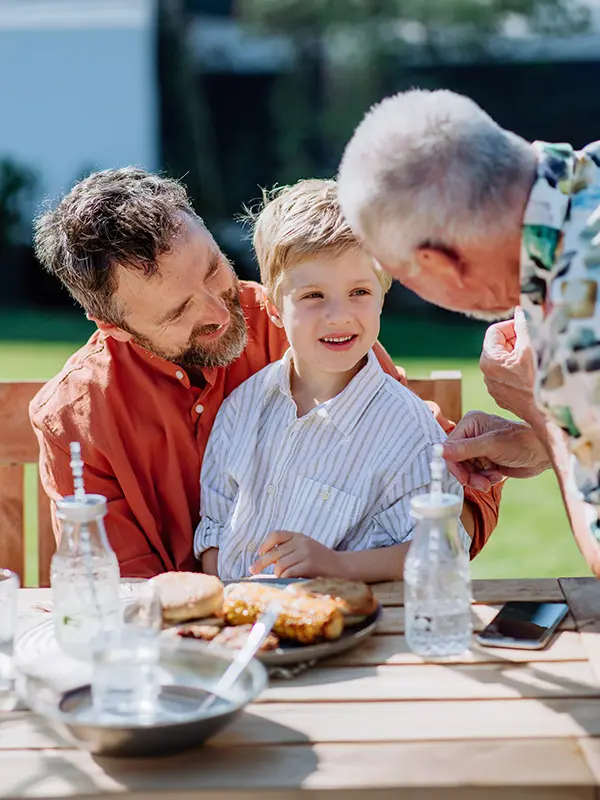 A young boy sits on a mans lap at an outdoor table, smiling at an older man leaning toward him. Food and drinks are on the table, and the scene is bright and cheerful, suggesting a family meal together.