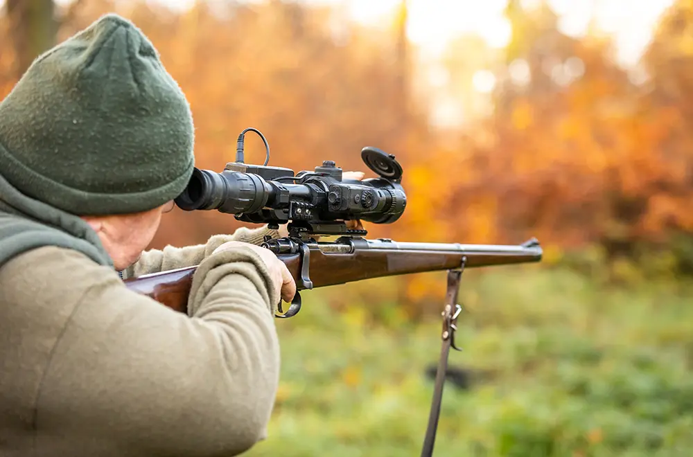 A person wearing a green beanie and jacket aims a rifle with a mounted scope in an outdoor setting with blurred autumn foliage in the background.