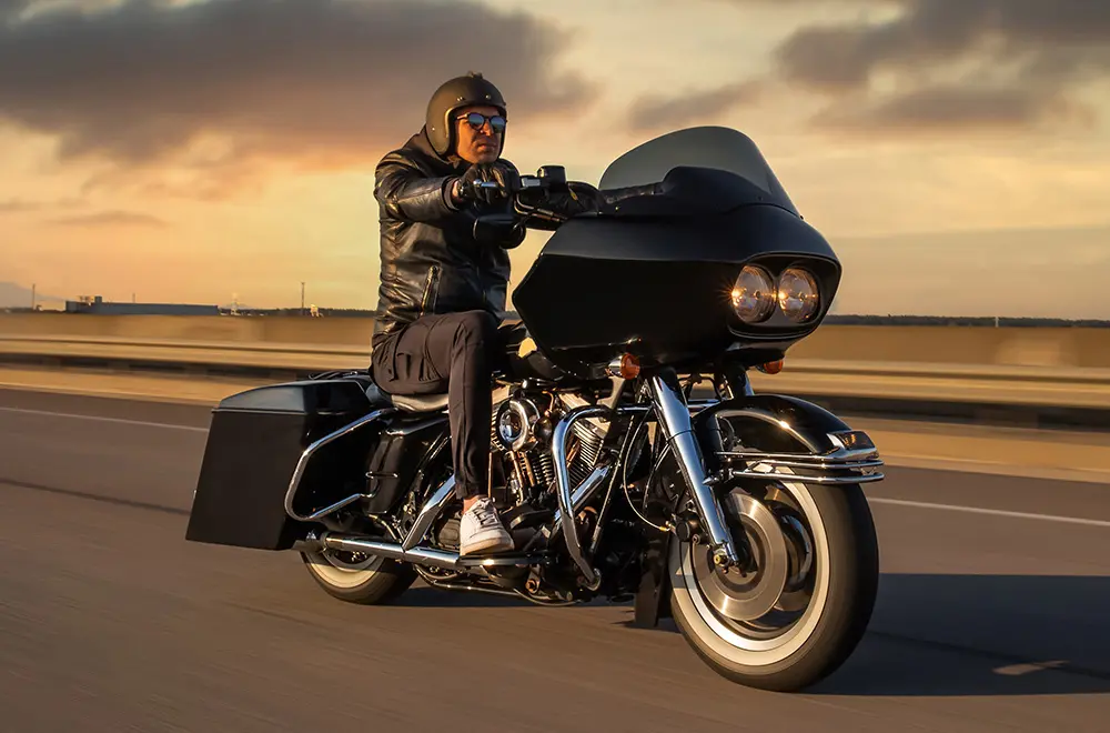 A person wearing a black helmet, sunglasses, and a leather jacket rides a large black touring motorcycle on a highway at sunset, with a blurred sky and road in the background.