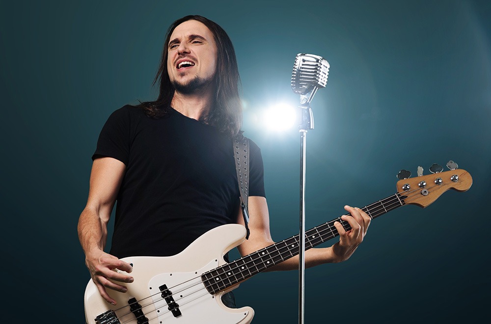 A man with long dark hair plays an electric bass guitar and sings passionately into a vintage microphone on stage, with bright stage lights shining behind him.