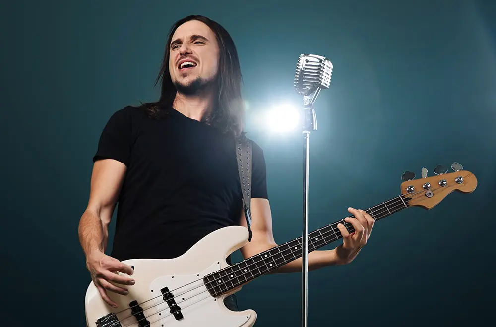 A man with long dark hair plays an electric bass guitar and sings passionately into a vintage microphone on stage, with bright stage lights shining behind him.