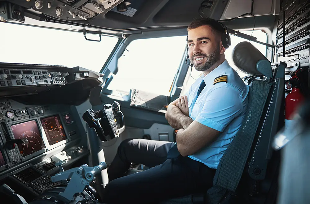 A male pilot in uniform sits in the cockpit of an airplane, smiling at the camera with his arms crossed. Cockpit controls, instruments, and windows are visible around him.