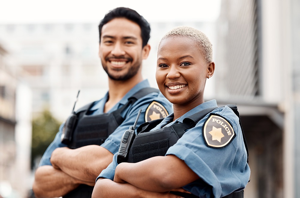 Two police officers, a man and a woman, stand side by side outdoors with their arms crossed, smiling at the camera. They wear police uniforms and radios, with blurred buildings in the background.