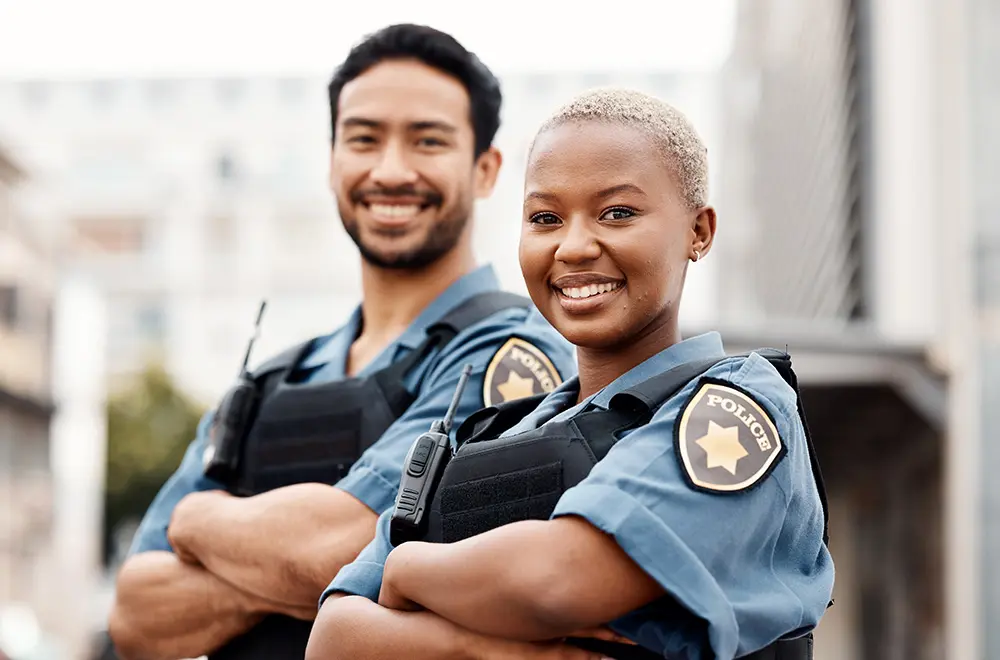 Two police officers, a man and a woman, stand side by side outdoors with their arms crossed, smiling at the camera. They wear police uniforms and radios, with blurred buildings in the background.
