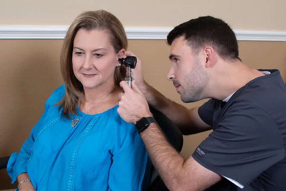 A healthcare professional examines a seated woman’s ear with an otoscope in a medical office. The woman wears a blue blouse and smiles slightly, while the professional focuses on the examination.