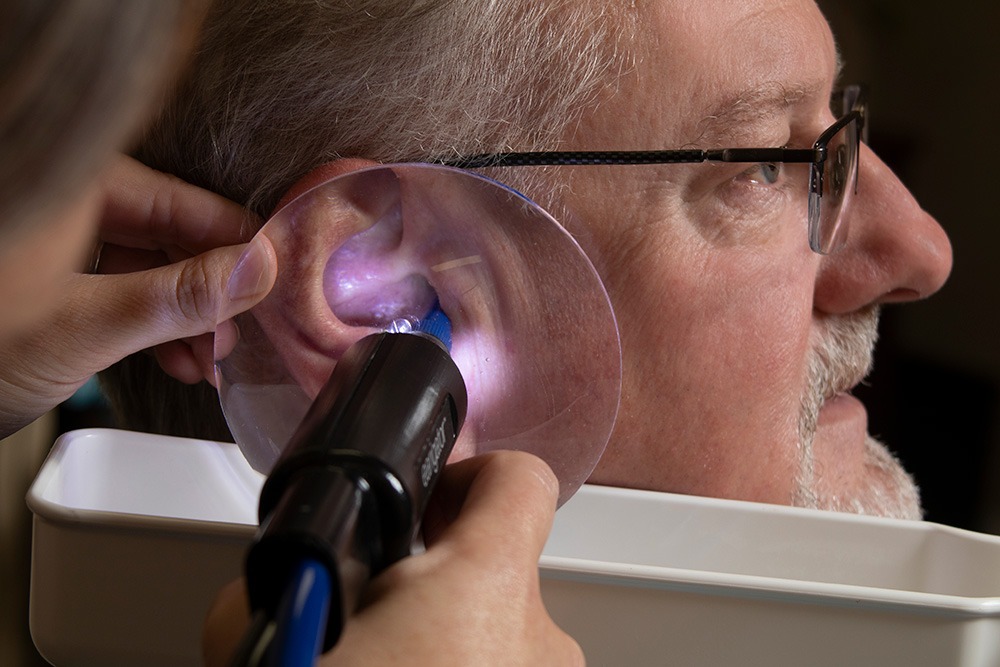 A close-up of an older man having his ear examined with a lighted tool and a clear plastic shield, likely during a professional ear cleaning or medical ear inspection.