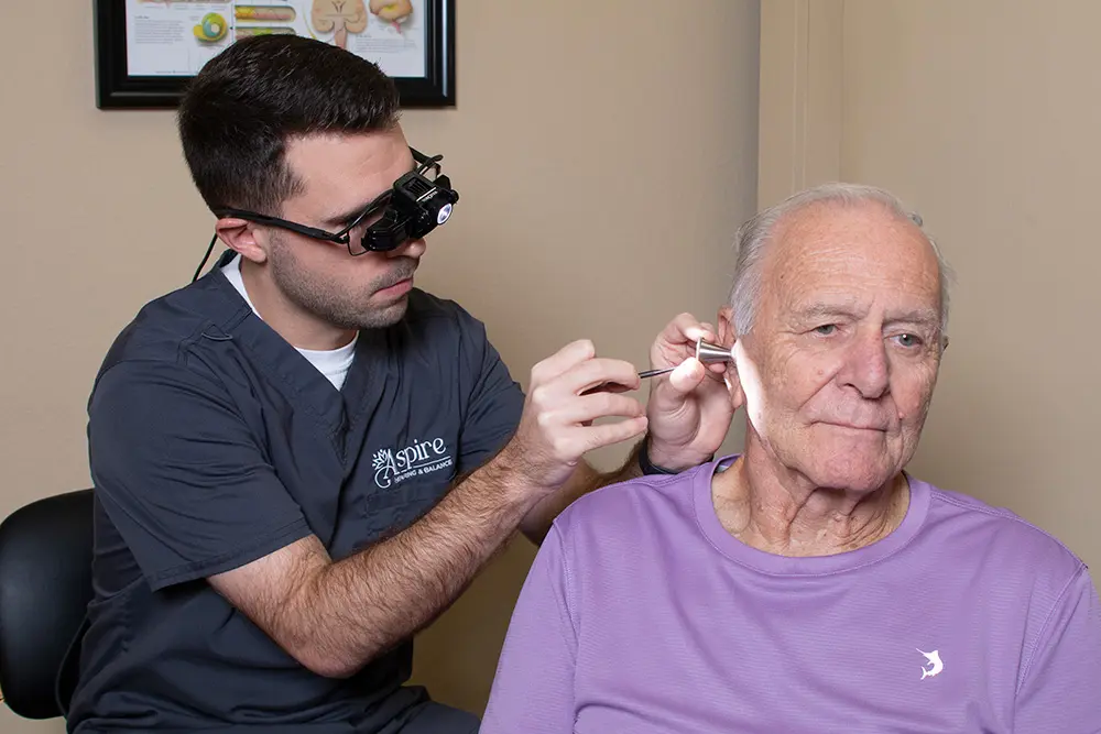 A healthcare professional uses a headlamp to examine an elderly mans ear with a medical tool. The man, wearing a purple shirt, sits calmly in a clinic room.