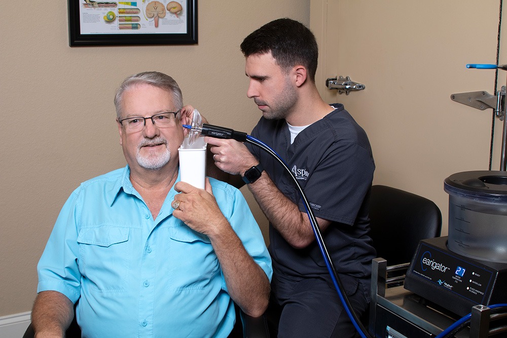 A healthcare professional in scrubs uses medical equipment on an older man’s ear in a clinic. The man sits and smiles, holding a cup under his ear. Medical charts and devices are visible in the background.