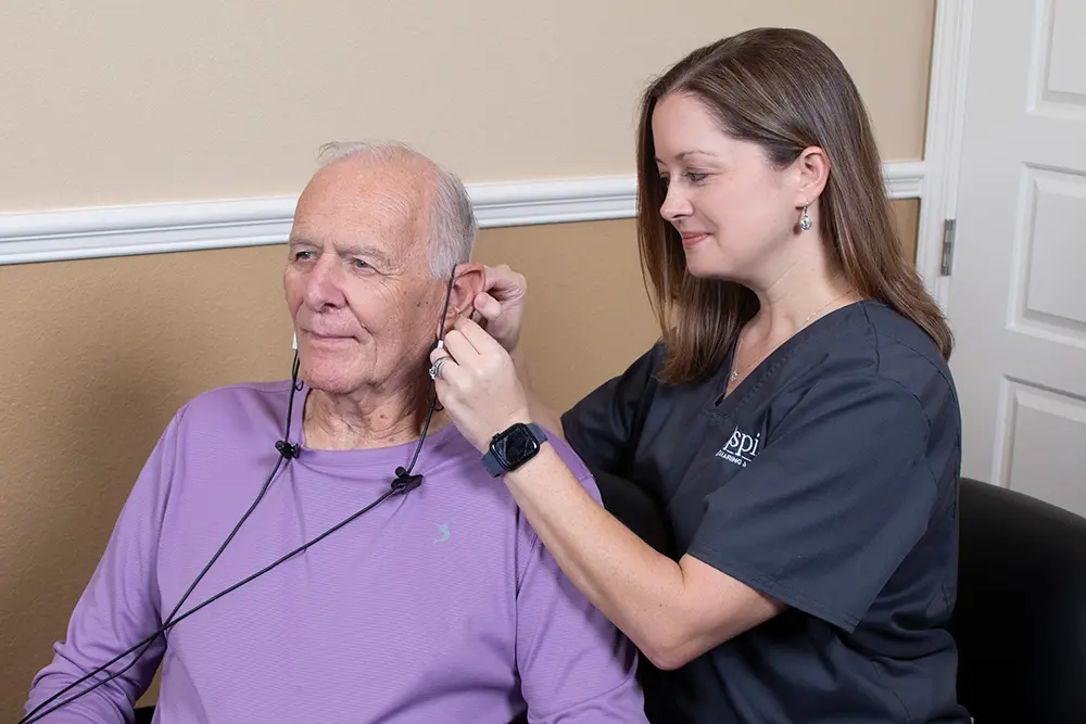 A woman in medical scrubs assists an older man by placing headphones or medical sensors over his ears in a clinical setting. The man is seated and smiling while the woman adjusts the equipment.