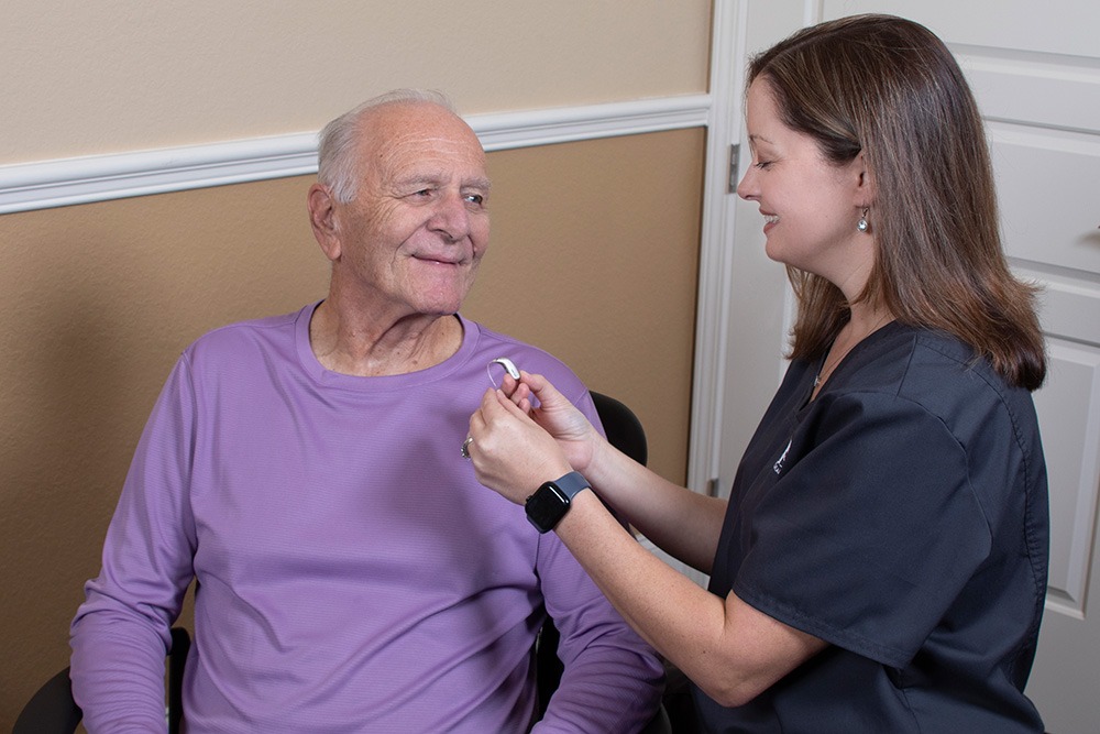 A female healthcare professional hands a hearing aid to an older man in a purple shirt, who is smiling while seated in a clinic setting.
