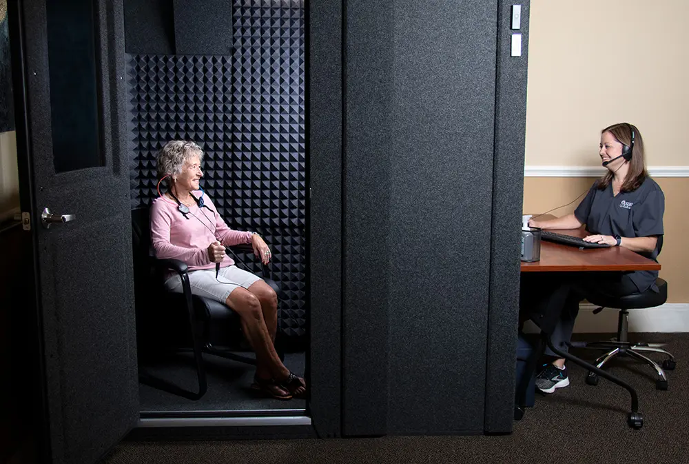 An older woman sits in a soundproof booth wearing headphones, undergoing a hearing test. A healthcare professional with a headset sits at a desk outside the booth, monitoring the test on a computer.