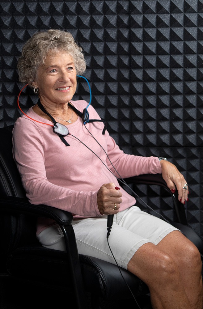An older woman sits in a soundproof room, wearing a headset with colored wires and holding a handheld device, likely undergoing a hearing or audio test. She is smiling and appears relaxed.