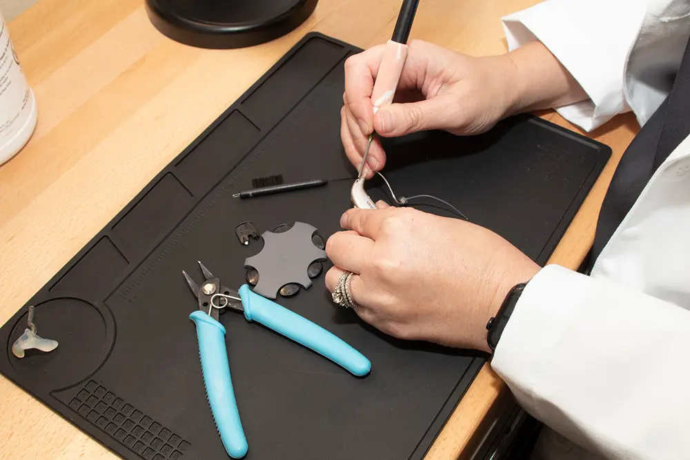A person wearing a white coat uses tools to adjust a hearing aid on a black work mat, with pliers, a cleaning brush, and a round gray tool nearby.