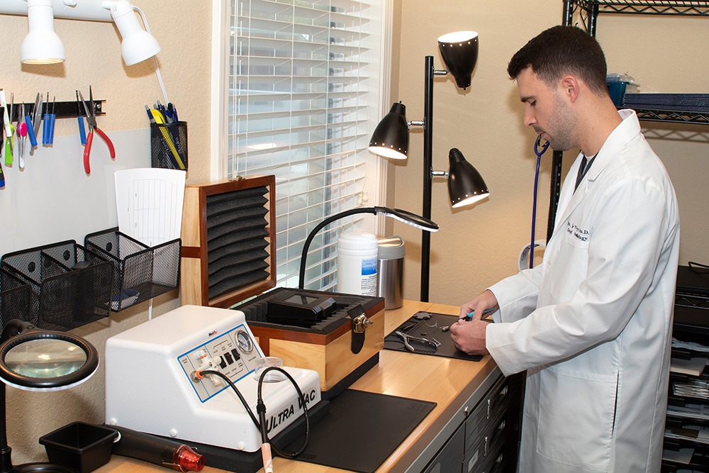 A man in a white lab coat works at a tool-filled workbench in a well-lit room. Various electronic equipment, tools, and lamps are organized on the counter and shelves beside him.