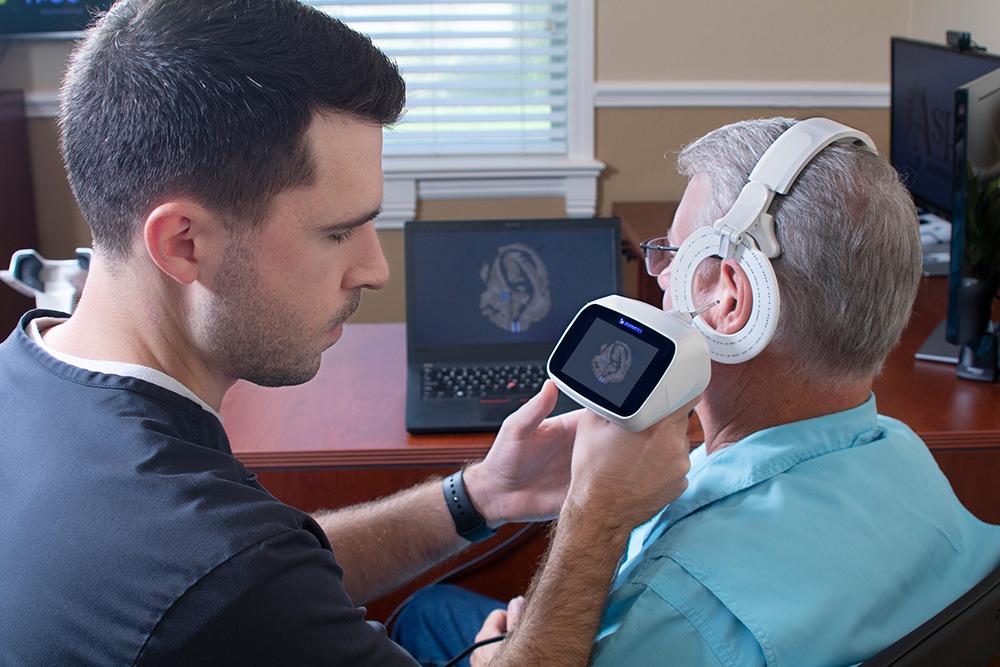 A healthcare professional uses a handheld medical device on an older mans ear while he wears headphones; a brain scan is visible on the device’s screen and a laptop in the background.