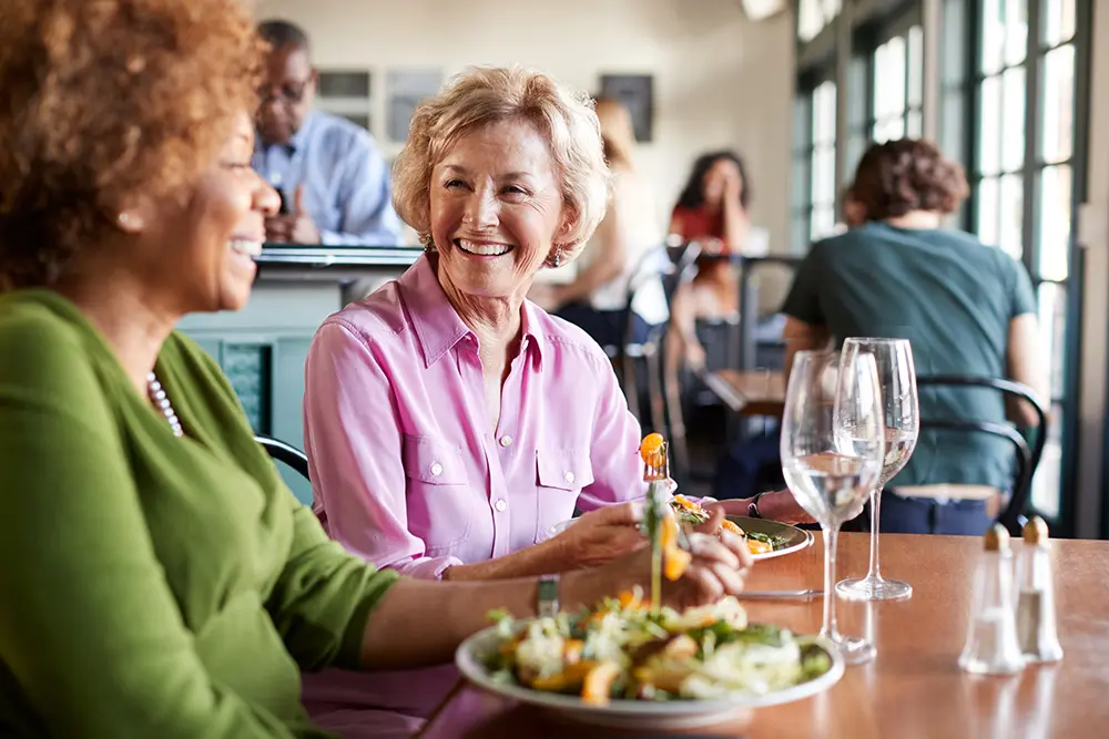 Two women sit at a restaurant table, smiling and enjoying a meal together. They have plates of salad and glasses of water in front of them, with other diners in the background.