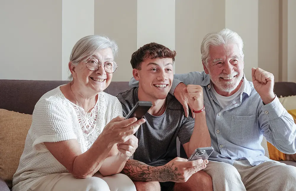 Three people, two seniors and a young adult, sit on a couch smiling and cheering while holding TV remotes, appearing happy and engaged, possibly watching a sports event or show together.