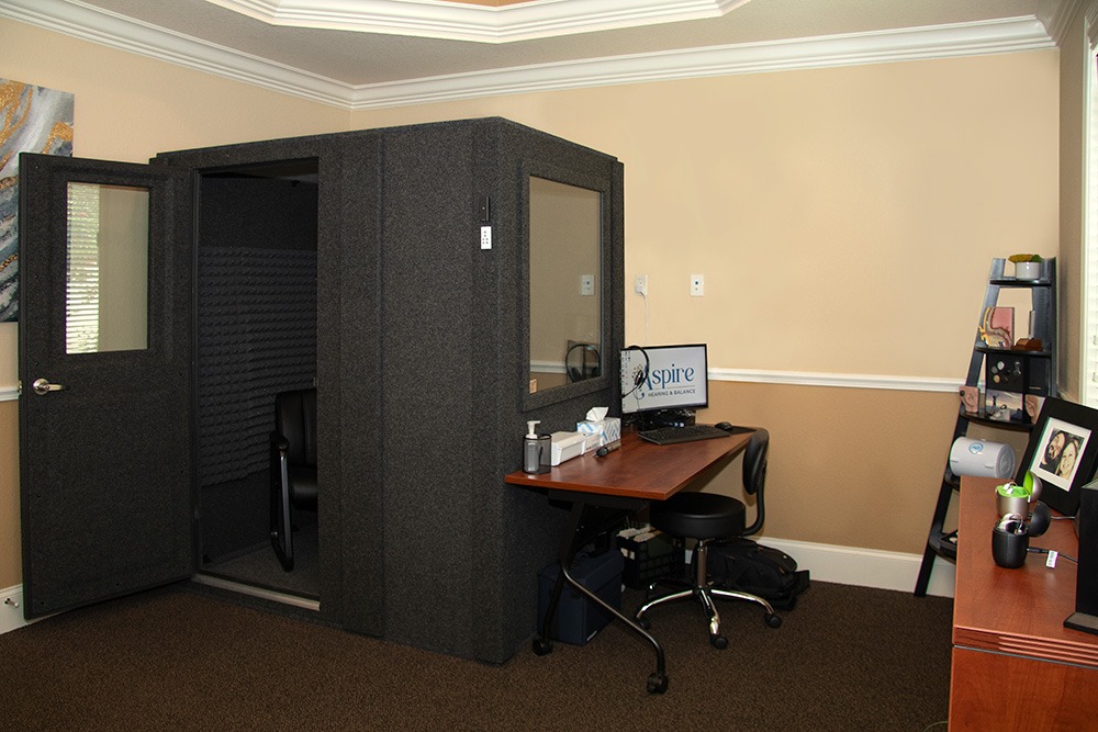 A small soundproof booth with an open door stands in a home office, next to a desk with a computer, office supplies, chair, and shelves with photos and decorations against beige walls and carpeted floor.