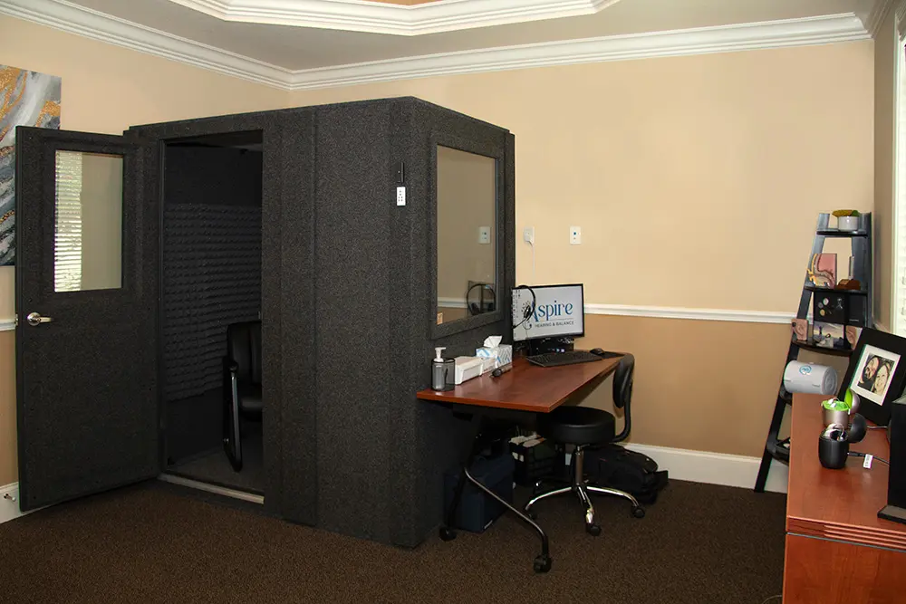 A small soundproof booth with an open door stands in a home office, next to a desk with a computer, office supplies, chair, and shelves with photos and decorations against beige walls and carpeted floor.