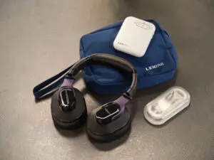 A Lenire device kit on a gray surface, including a pair of headphones, a white handheld device, a blue carrying pouch, and a small clear case with white earpieces inside.