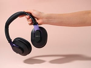 A hand holds black over-ear headphones with purple accents against a light pink background.