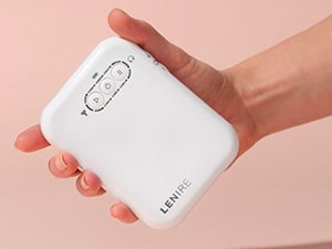 A hand holding a white LENIRE device with three buttons on the front, set against a soft pink background.