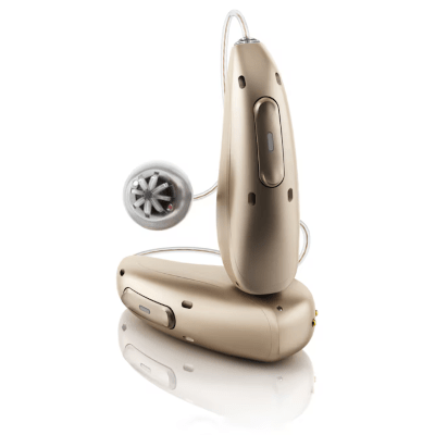 Two beige behind-the-ear hearing aids are shown; one is upright, and the other is lying down. The upright device has a clear wire ending in a round, perforated earbud. The devices are reflected on a white surface.