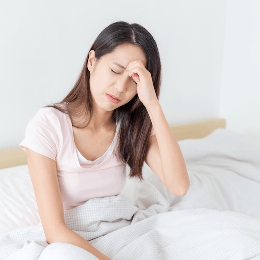 A woman sitting in bed with her eyes closed, holding her forehead and appearing to be in pain or discomfort.
