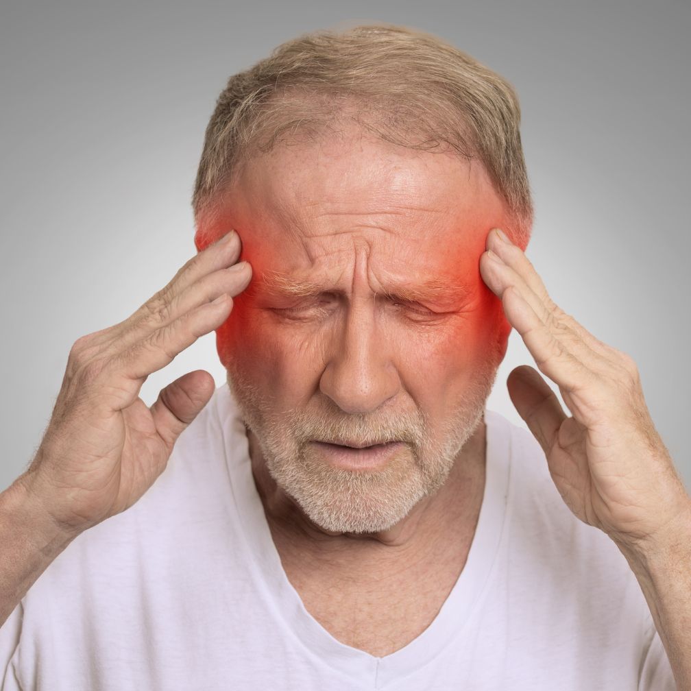 An older man with closed eyes holds his temples, showing discomfort or pain. Red highlights on his temples suggest a headache or migraine.