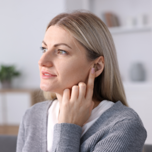 A woman with long blonde hair, wearing a gray sweater, touches her ear with her finger. She looks thoughtful and is gazing slightly upward. The background is softly blurred with home decor.