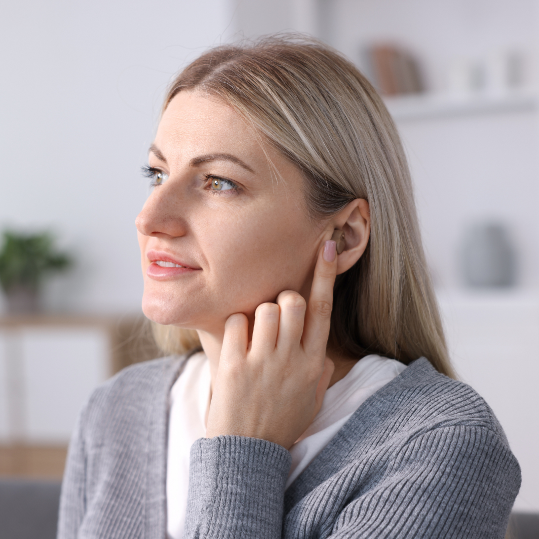 A woman with long blonde hair, wearing a gray sweater, touches her ear with her finger. She looks thoughtful and is gazing slightly upward. The background is softly blurred with home decor.