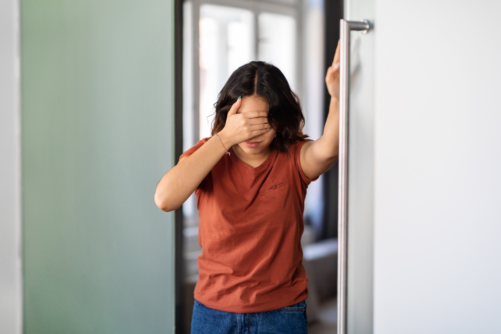 A woman in an orange t-shirt covering her eyes with her hand while leaning against a door frame.