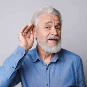 An elderly man with a white beard wearing a blue button-down shirt cups his hand behind his ear to listen.